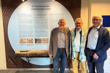 Drei Personen stehen vor der Ausstellung „Ein Teuchel aus dem heilsamen Bad zu Boll“ im Kurhaus Bad Boll. Im Vordergrund ist der historische Teuchel auf einer von Stahlbau Nägele gefertigten Rundstahl-Unterkonstruktion zu sehen.