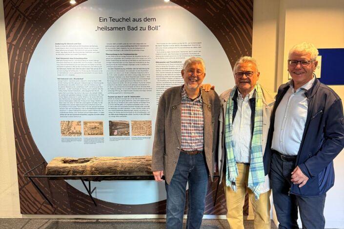 Drei Personen stehen vor der Ausstellung „Ein Teuchel aus dem heilsamen Bad zu Boll“ im Kurhaus Bad Boll. Im Vordergrund ist der historische Teuchel auf einer von Stahlbau Nägele gefertigten Rundstahl-Unterkonstruktion zu sehen.