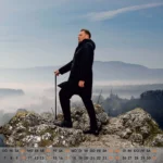 Tableau vivant zum Wanderer über dem Nebelmeer, Mann in schwarzem Mantel steht mit Stock auf Felsen und blickt über neblige Landschaft und Berge