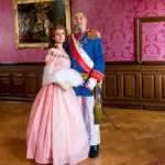Tableau vivant Das Kaiserpaar, Frau im rosa Ballkleid mit Fächer steht neben Mann in blauer Uniform mit Schärpe und Orden, beide in prunkvollem Salon