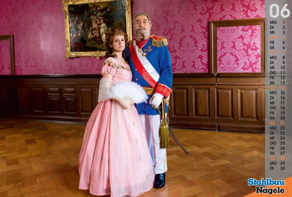 Tableau vivant Das Kaiserpaar, Frau im rosa Ballkleid mit Fächer steht neben Mann in blauer Uniform mit Schärpe und Orden, beide in prunkvollem Salon